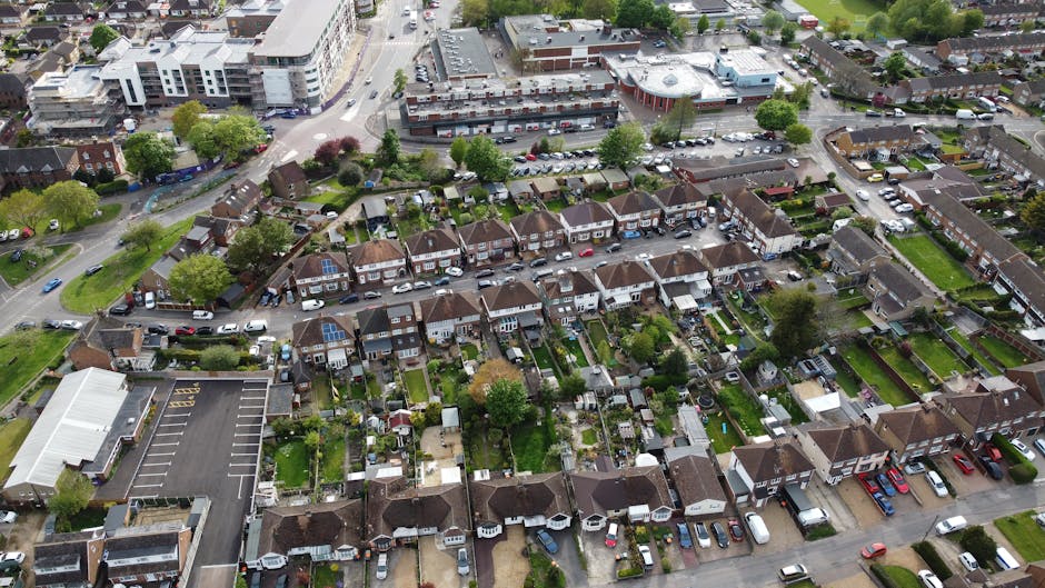 An aerial view of a residential neighborhood showcasing a row of terraced houses with small private gardens, featuring lush green lawns, various trees, and well-maintained flower beds. Some gardens ha