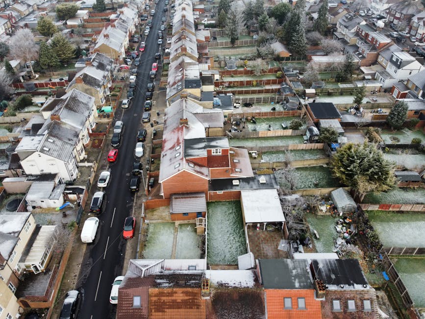 An aerial view of a residential neighbourhood showing tightly packed terraced houses with pitched roofs, some of which have patches of snow. The street runs centrally through the image, lined with par